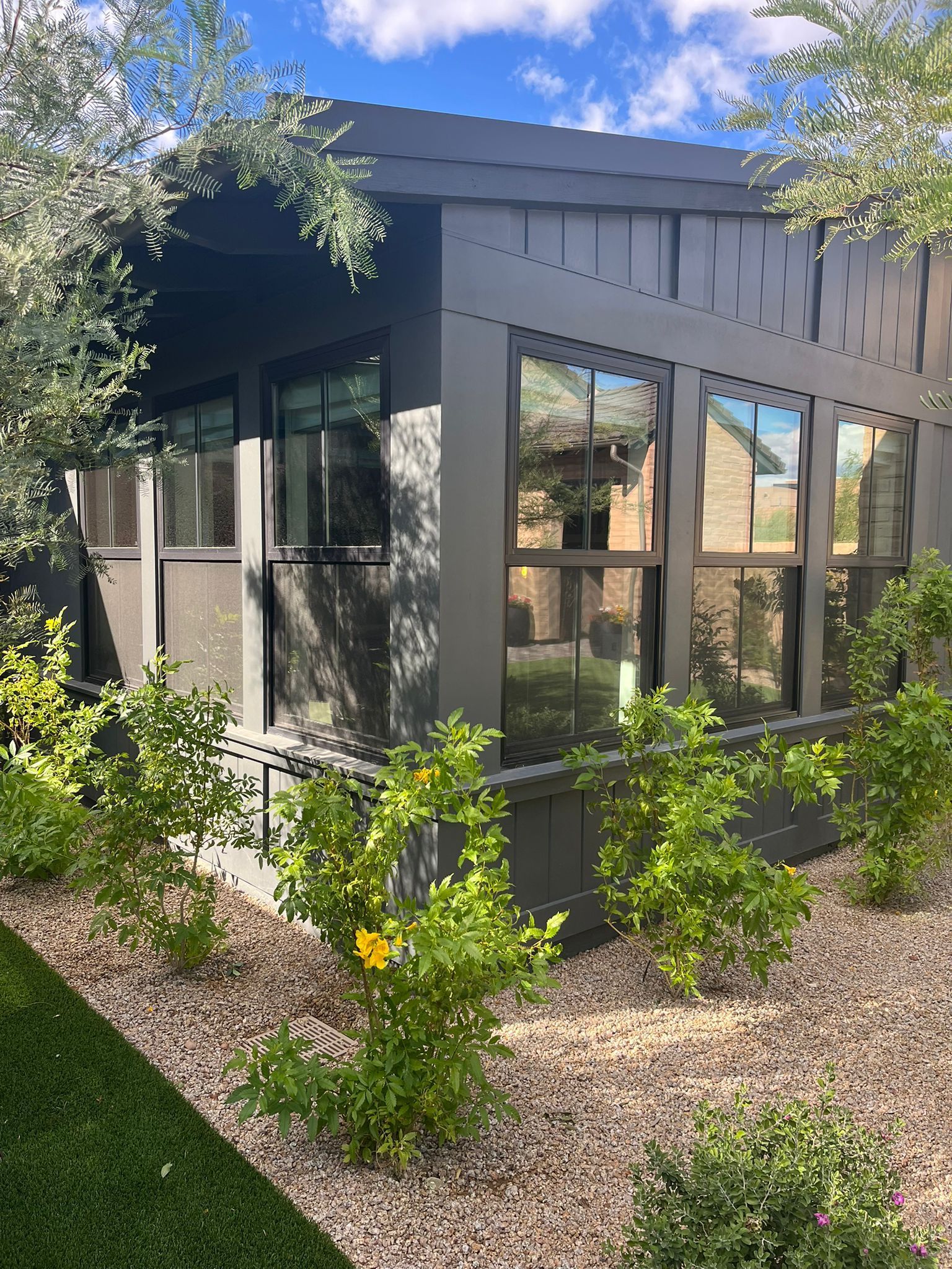 Modern gray house exterior with large windows, surrounded by desert landscaping and green shrubs.
