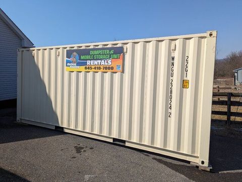 White shipping container with a rental sign, parked on asphalt beside a fence and building.