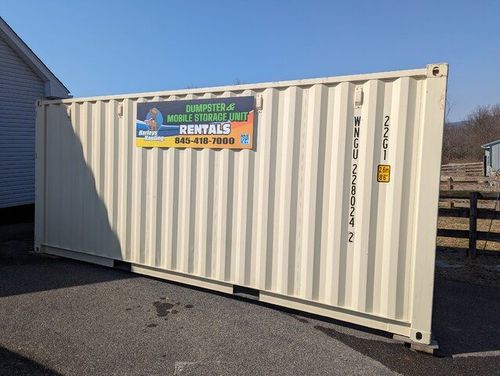 White shipping container with a rental sign, parked on asphalt beside a fence and building.
