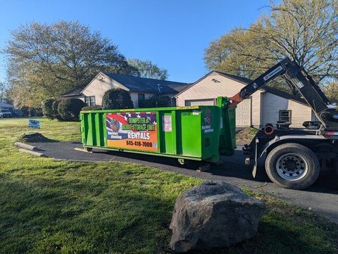 Green dumpster on a residential driveway beside a house, with a truck attached and a large boulder in front.