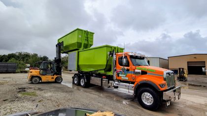 Orange truck carrying a green dumpster with a forklift nearby in a muddy industrial lot