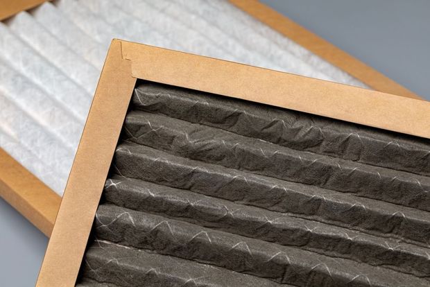 Black pleated air filter in a tan cardboard frame on a white ribbed surface