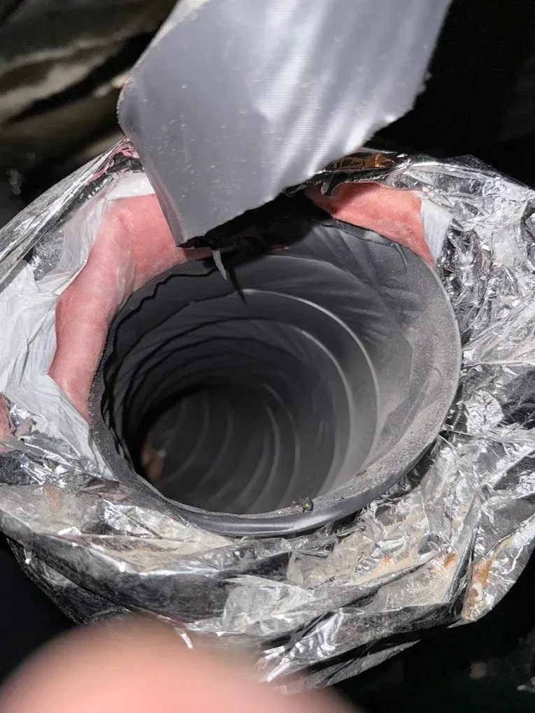 Close-up of a black flexible duct opening wrapped in clear plastic, with a gray flap lifted back.