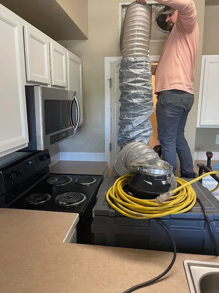 Person installing a large flexible air duct in a kitchen, with a vacuum machine and coiled hose nearby.