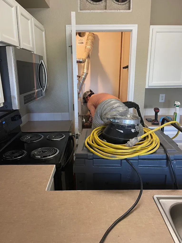Worker in kitchen crawlspace with vacuum hose and coiled yellow cord, repairing or cleaning behind cabinets.