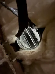 Close-up of a circular metal vent or duct seen through insulation inside a wall cavity.