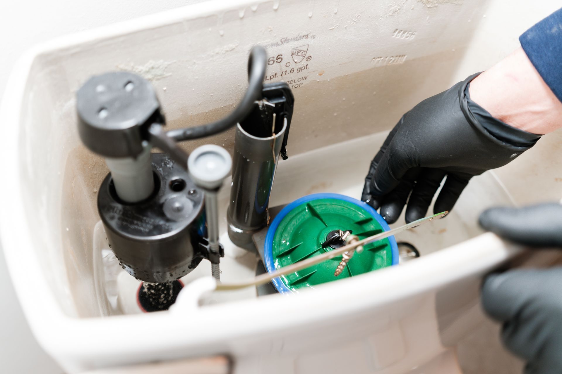 Gloved hands repairing a toilet tank with exposed plumbing parts and tools inside.