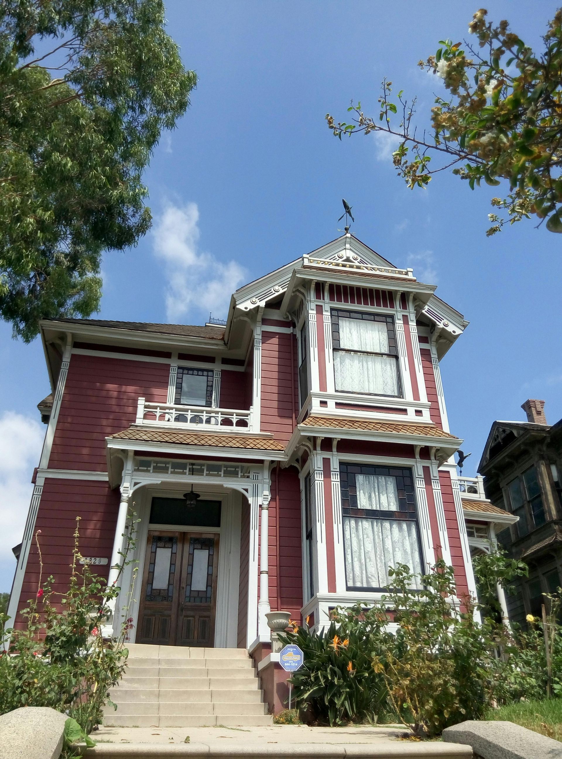 Victorian-style red house with ornate white trim, front steps, and garden under a blue sky