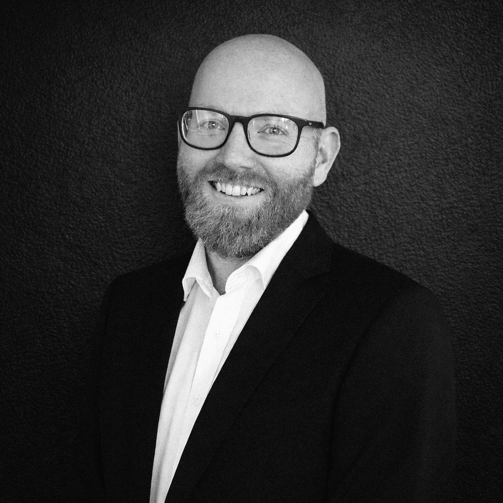 Bald man with glasses, red beard, smiling, wearing a suit jacket and white shirt against a gray backdrop.