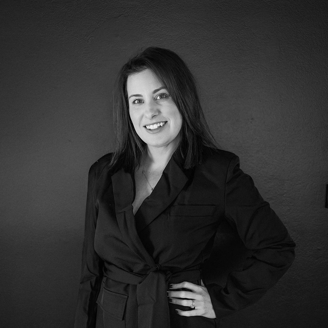 Woman in a black blazer smiles, hand on hip. Dark background.
