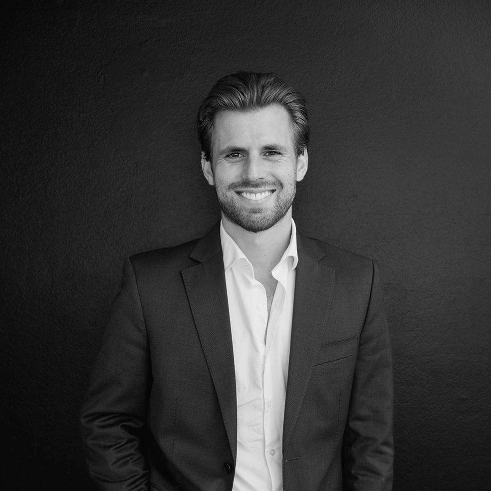Man in a suit smiling, leaning against a dark wall.