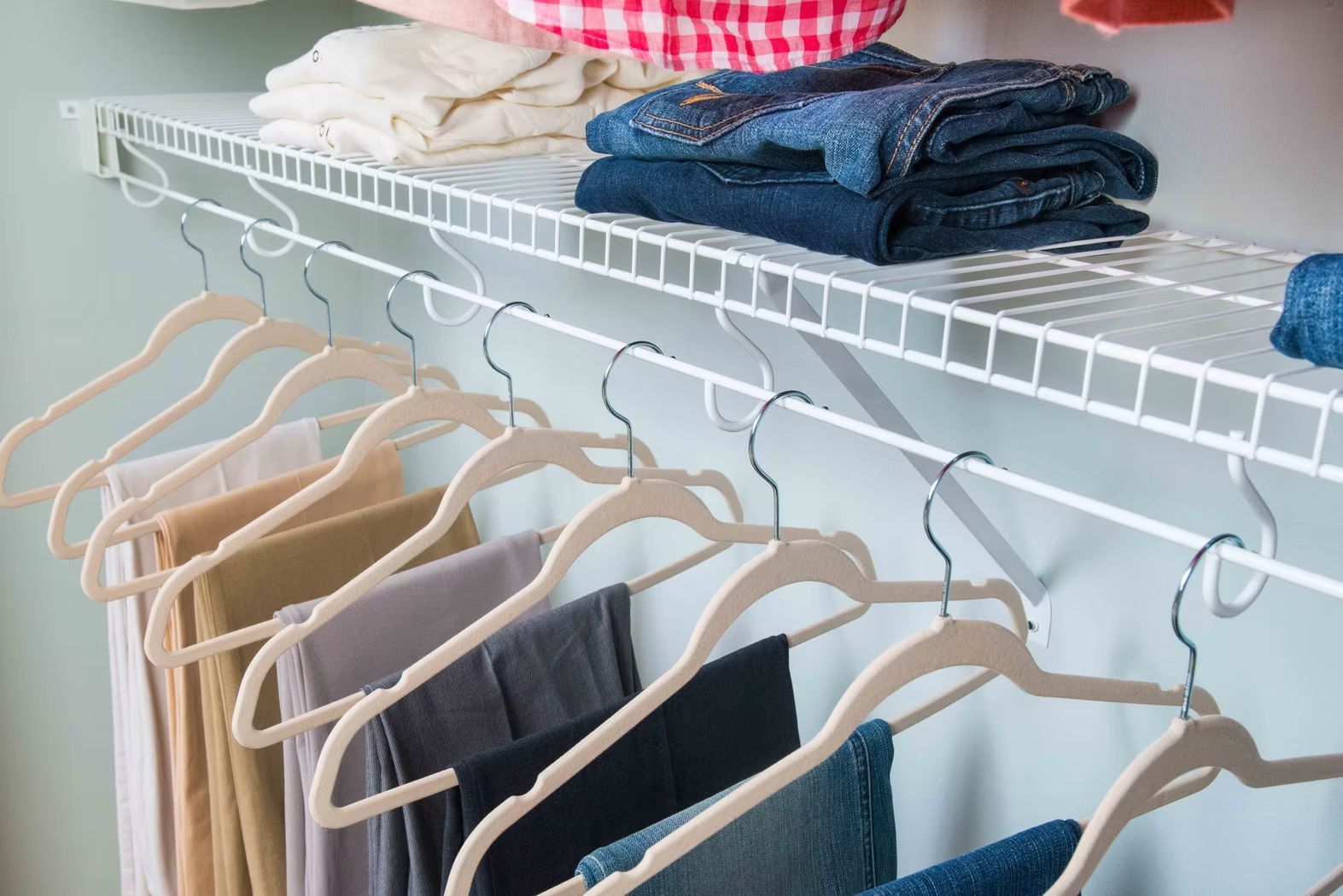 Clothes hanging on a white wire closet rack with folded jeans and shirts on the shelf above