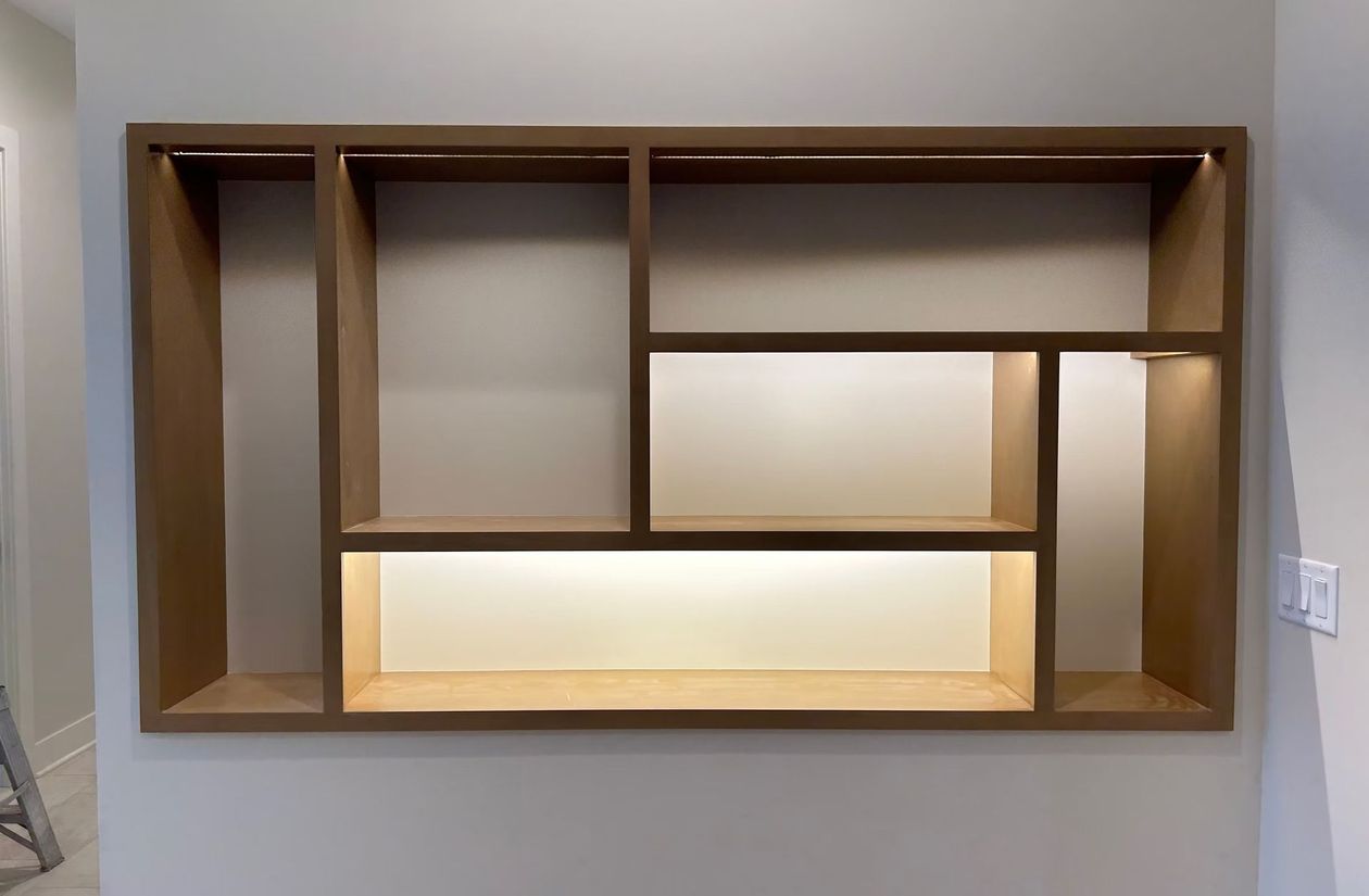 Wooden wall-mounted geometric shelving unit on a light-colored wall with backlit central compartments