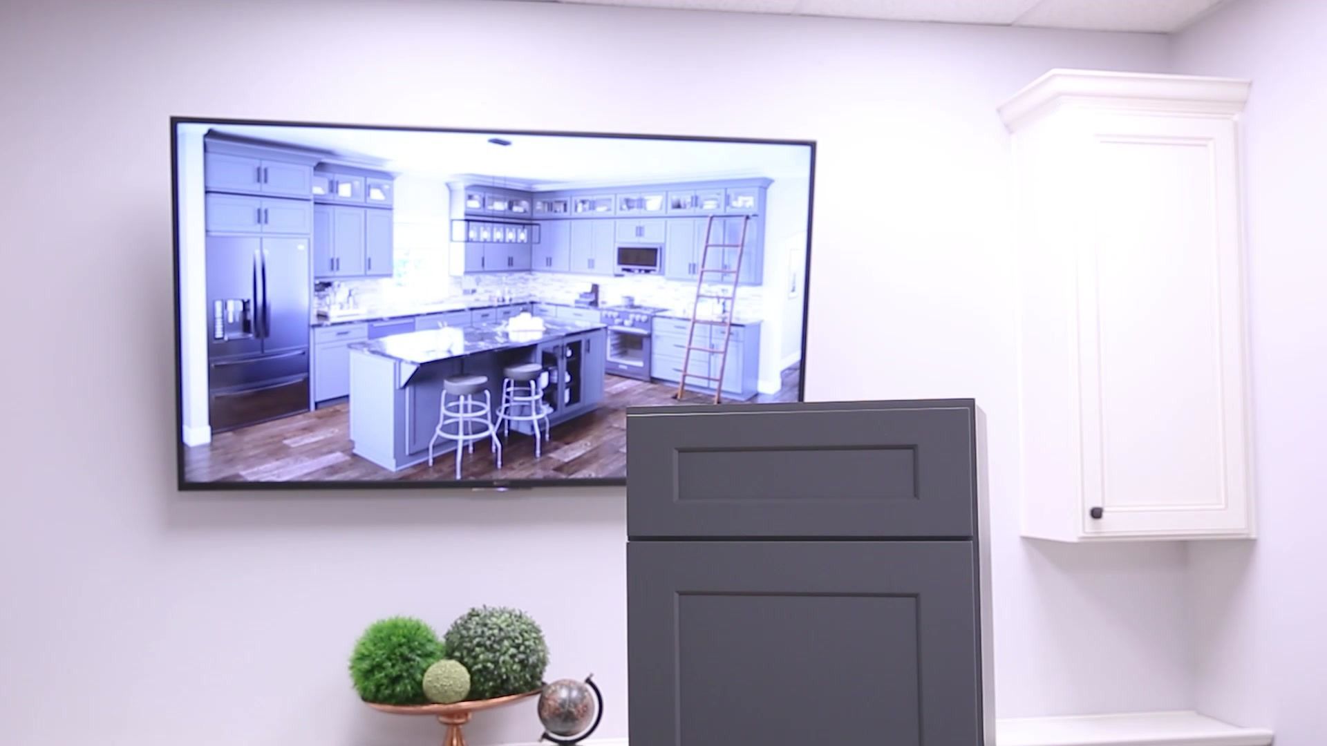 Wall-mounted TV showing a kitchen, with a cabinet and decor in a bright modern room.