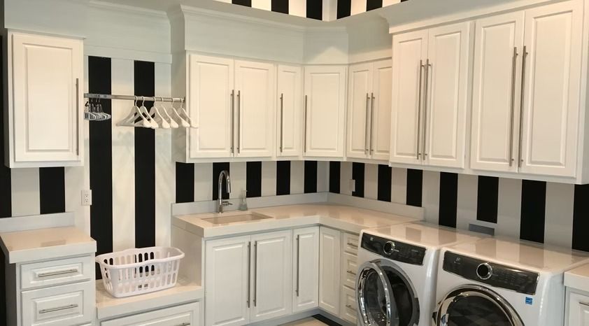 White L-shaped laundry room with cabinets, shelves, hanging rod, and washer-dryer under black-and-white striped walls