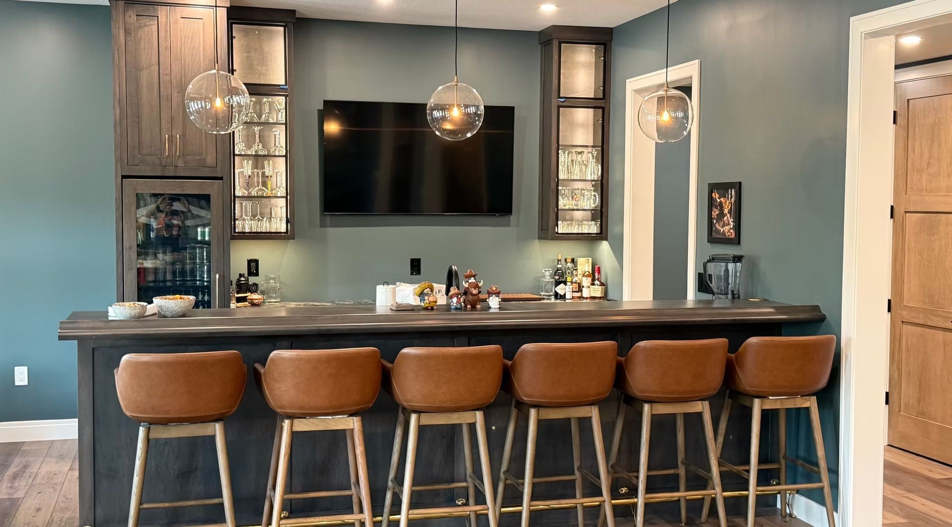 Modern home bar with five brown stools, dark cabinetry, wall-mounted TV, and pendant lighting.