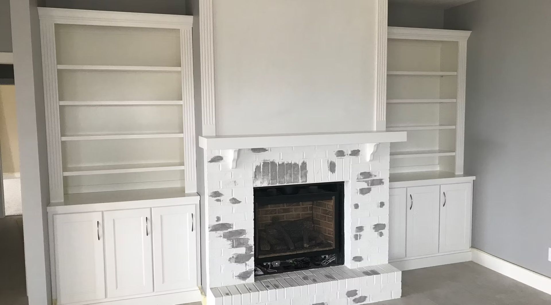 Built-in white shelves and cabinets flanking a white fireplace with a dark firebox in a gray room