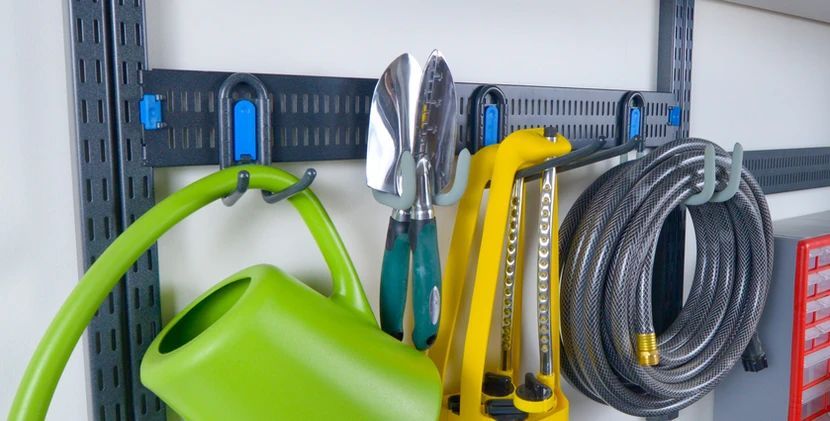 Green watering can, silver tools, yellow hose, and coiled gray hose hanging on a wall rack.