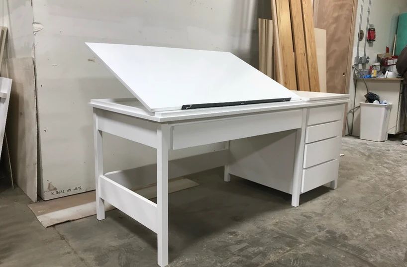 White drafting table with an angled top and drawers in a workshop, with wood planks nearby