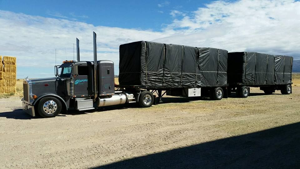 Black semi-truck hauling two covered trailers on a dirt lot under a blue sky
