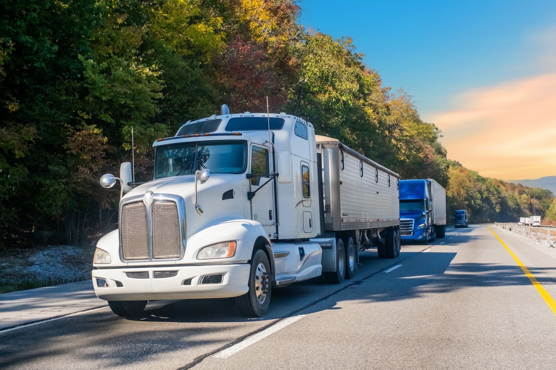 White semi-truck hauling a trailer on a highway, with a blue truck behind and trees lining the road.