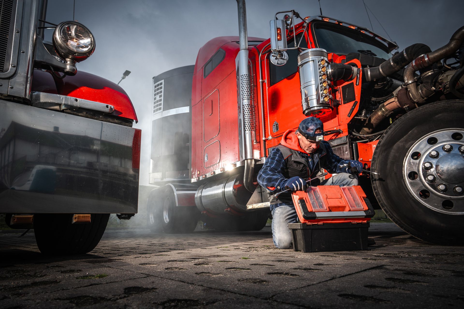 Mechanic crouches by a red semi-truck in a garage, with another truck parked nearby.