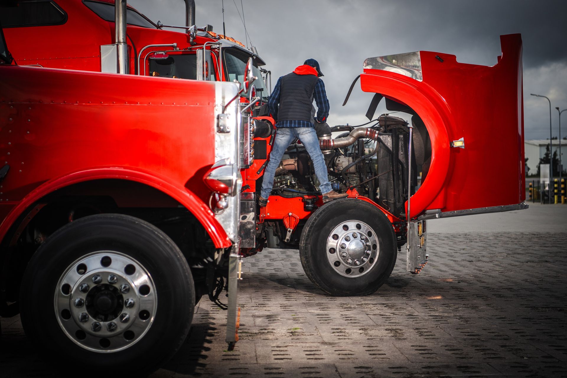 Person working on a red truck with its hood open in a cobblestone lot
