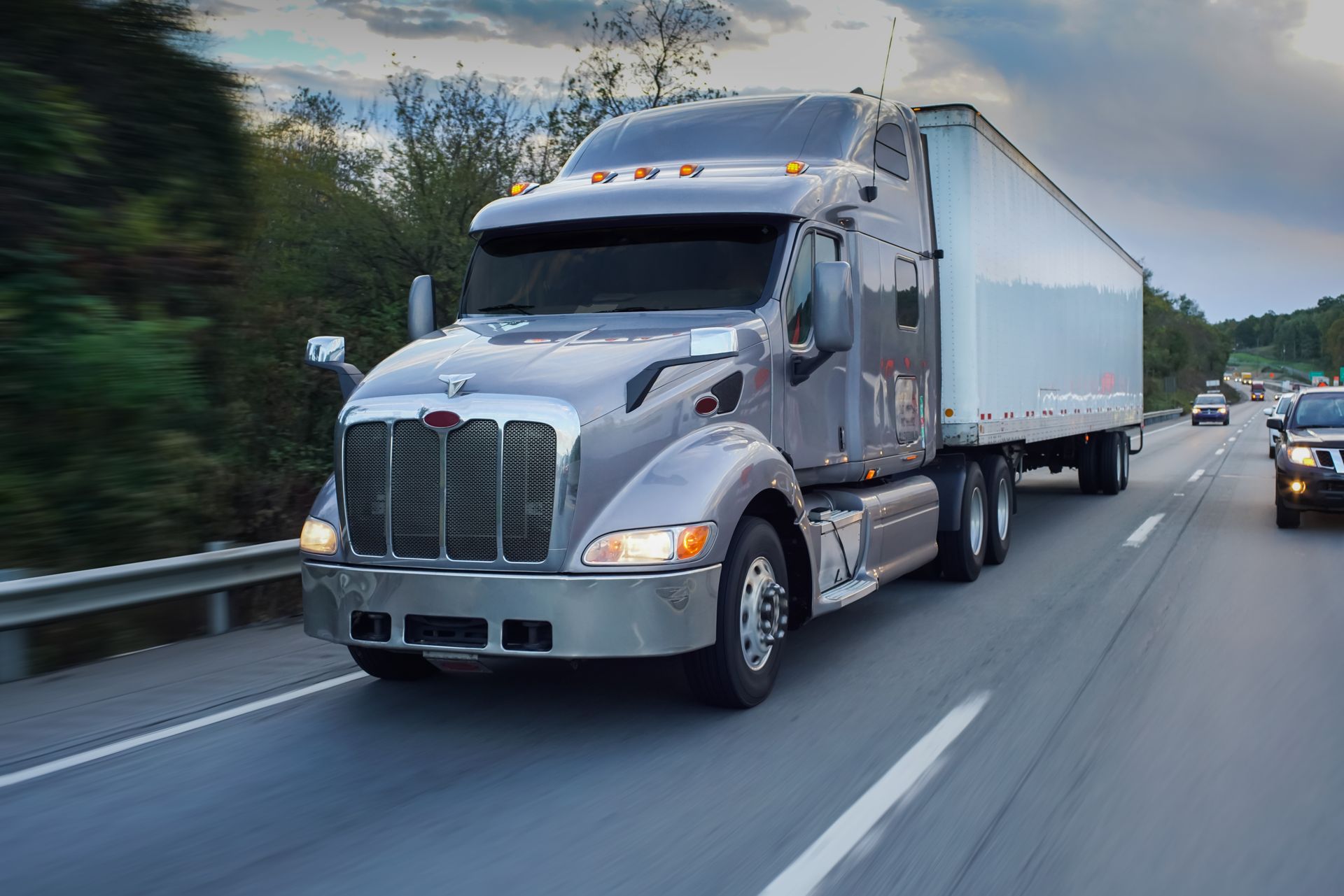 Silver semi truck driving on a highway beside trees and cars