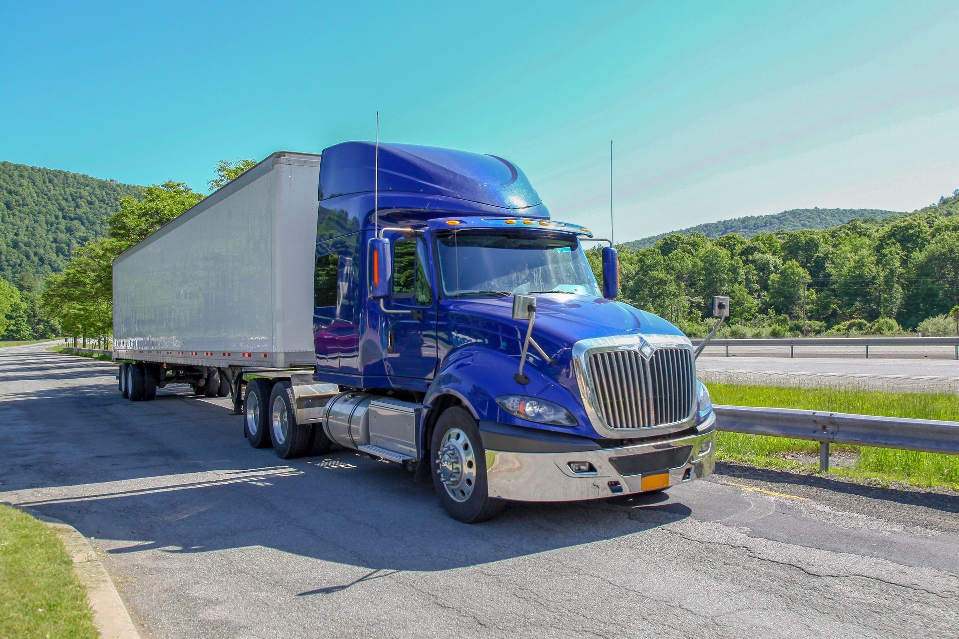 Blue semi-truck driving on a sunny highway with green hills in the background