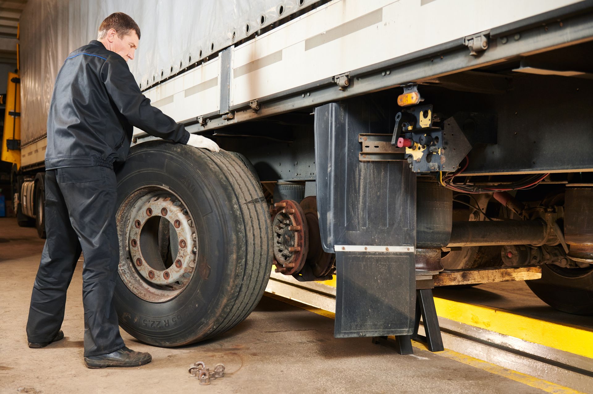 Mechanic inspecting a truck’s rear wheel under a raised trailer in a garage