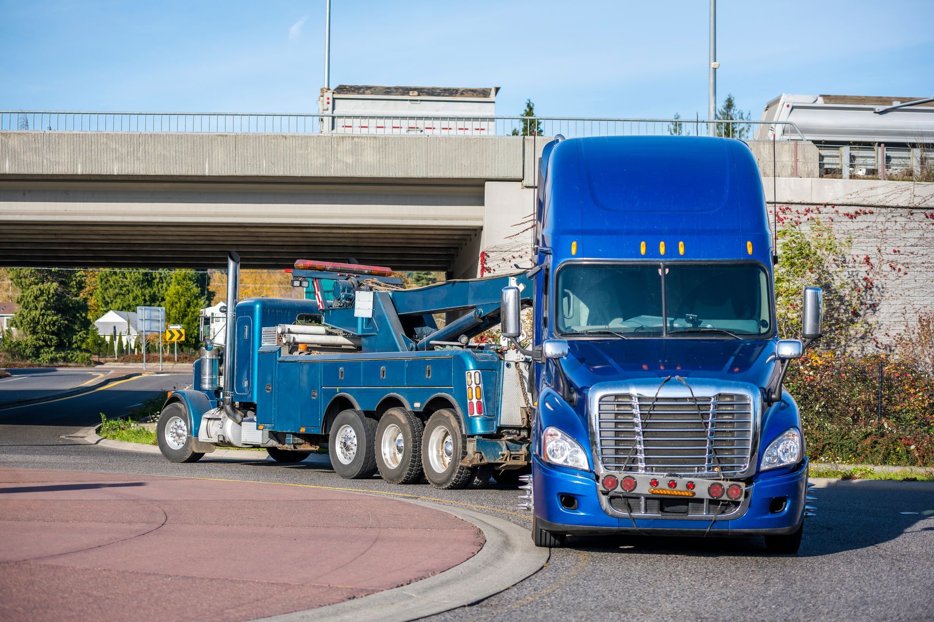 Blue semi-truck and tow truck under a highway overpass on a roadside curve
