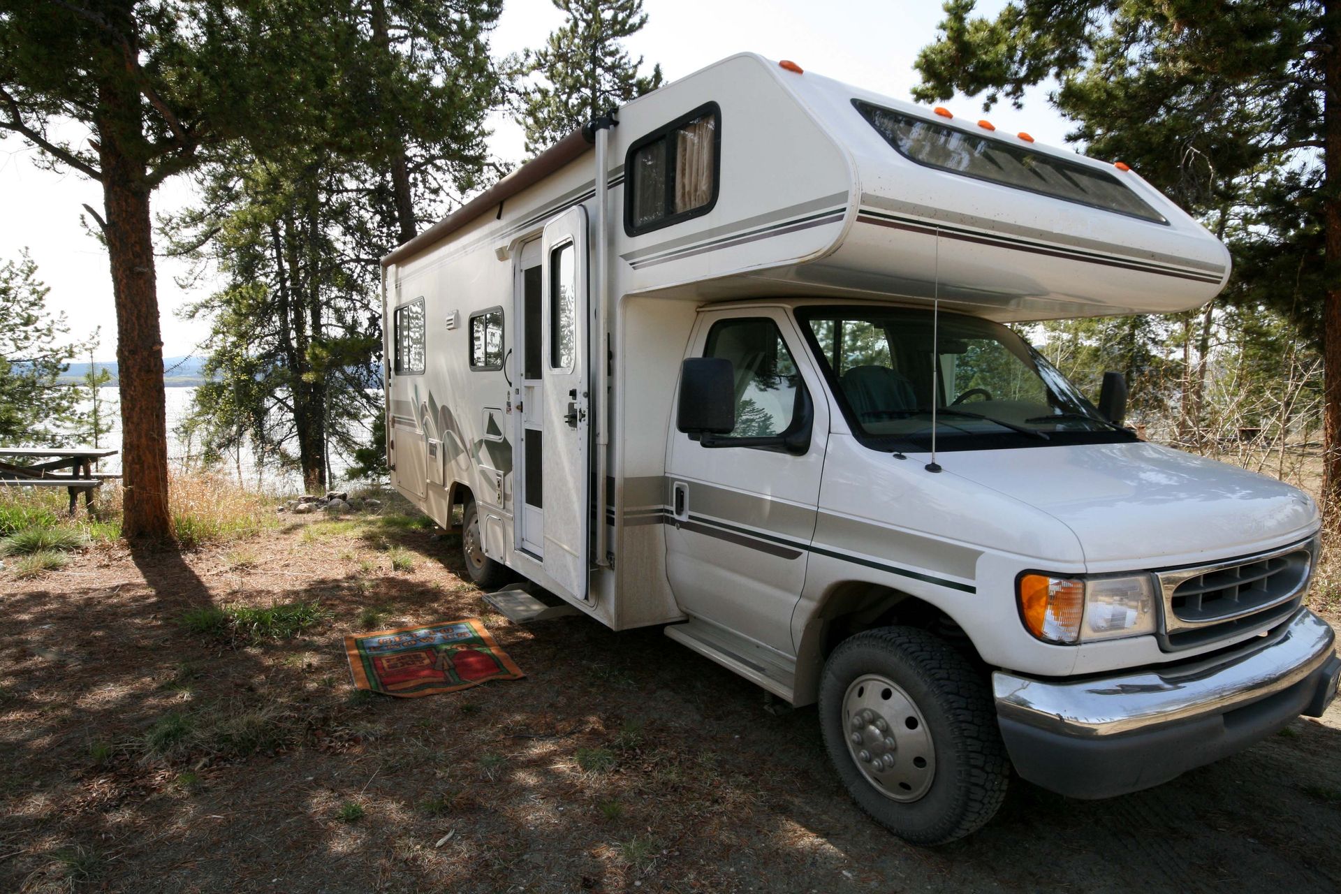 White RV parked on a wooded dirt campsite with trees and sunlight.