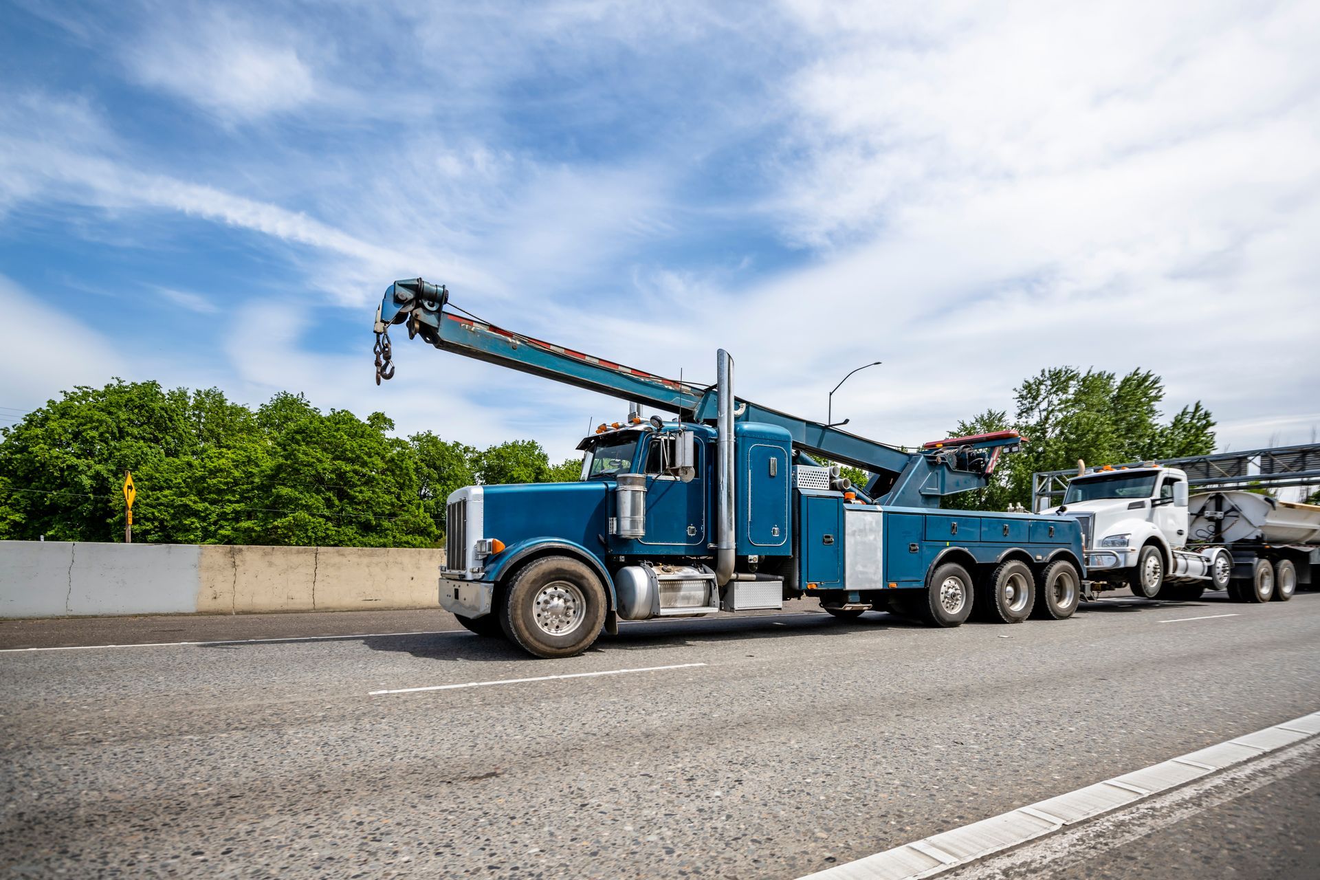 Blue tow truck towing a white truck on a road under a cloudy sky