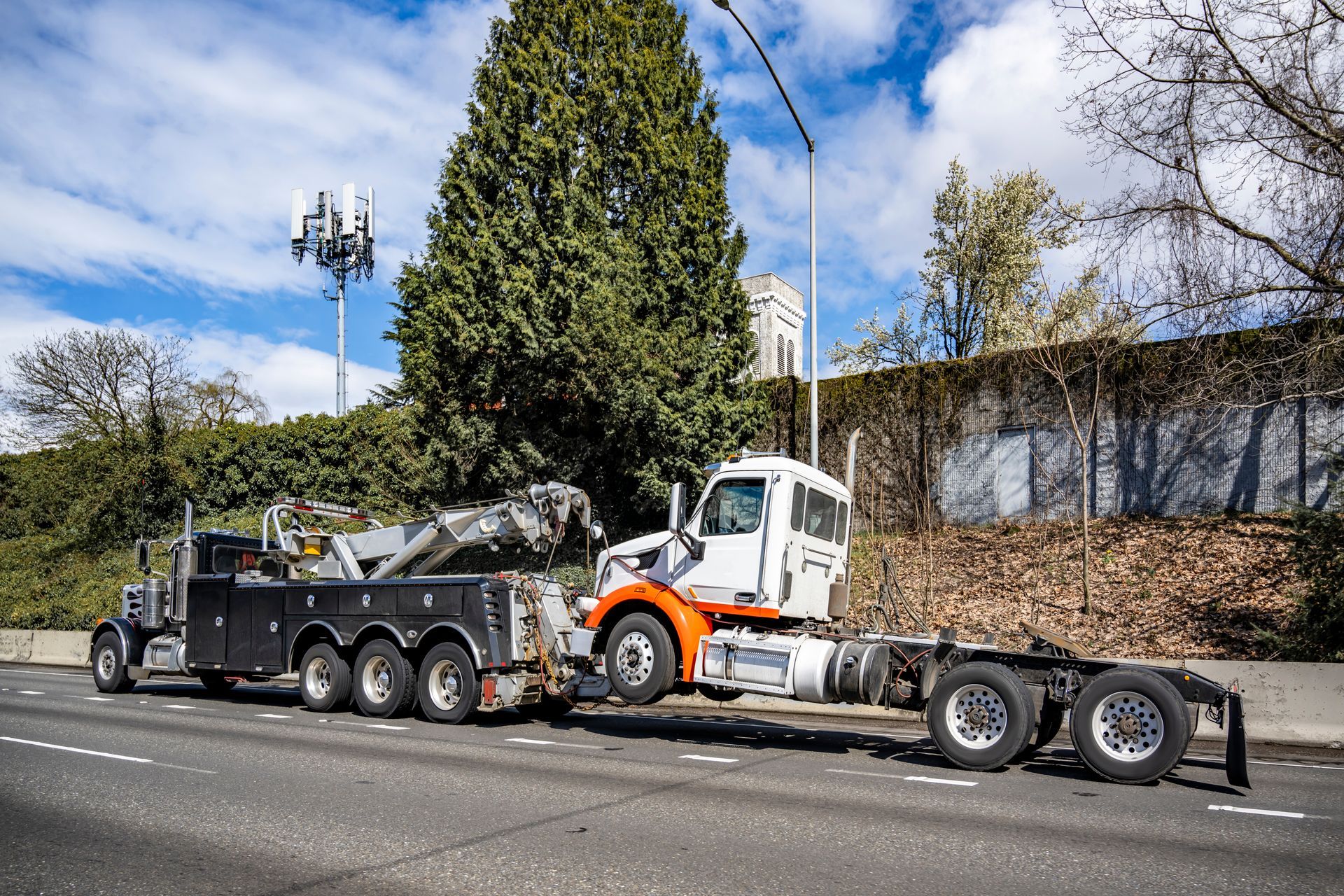 Tow truck hauling a damaged dump truck along a roadside, with trees and a cell tower behind it