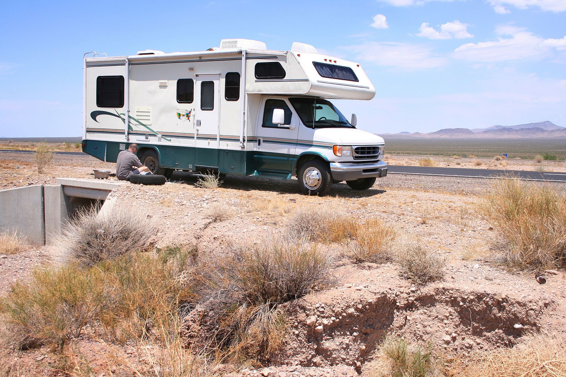 White RV parked on a dirt road in a desert landscape with sparse shrubs and distant mountains.