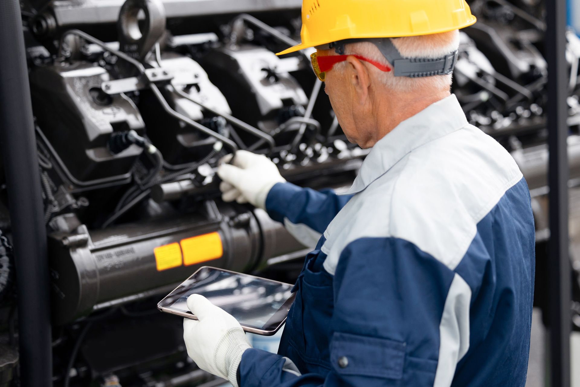 Technician in hard hat and gloves inspecting industrial machinery with a clipboard in a factory