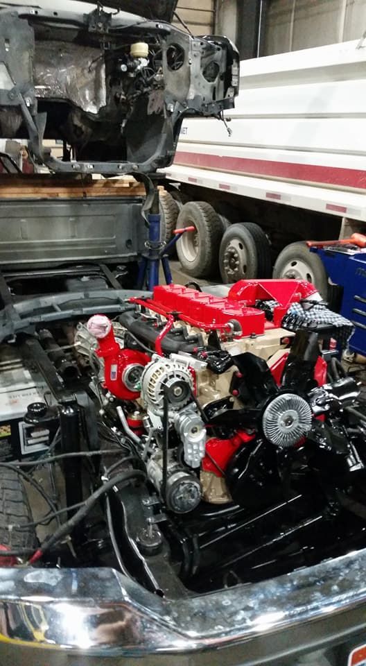 Red engine in a car chassis at a repair shop, with a truck and workshop equipment in the background