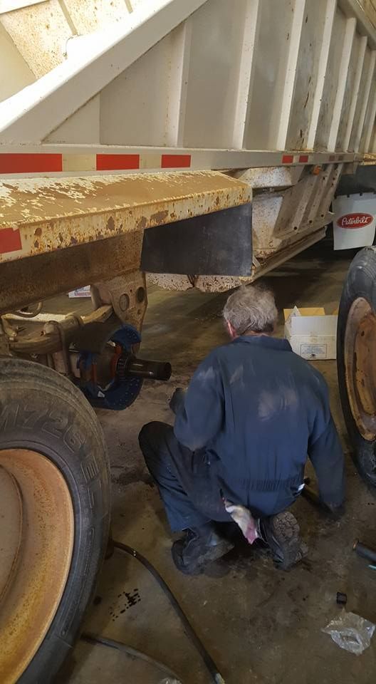Worker kneeling under a dump truck, inspecting the underside near the rear wheels in a muddy work area.