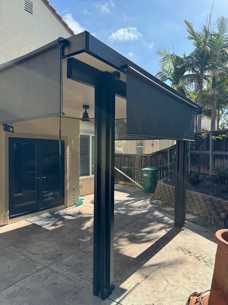 Backyard patio with a black metal pergola attached to a house, on a sunlit paved area