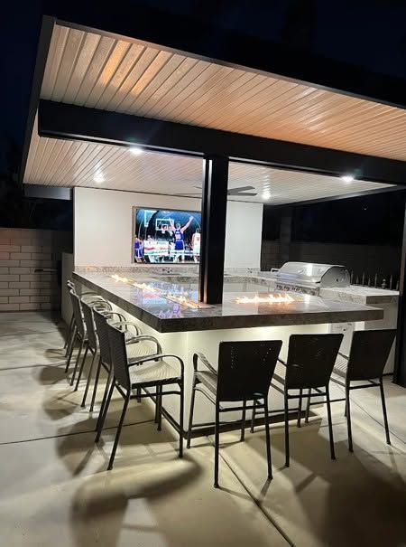 Covered outdoor patio with a long stone table, bar stools, chairs, and a TV under warm lighting.