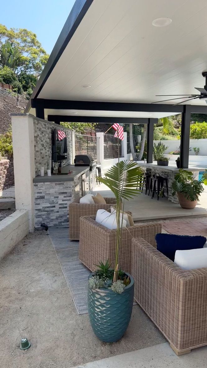Covered outdoor patio with wicker seating, potted plants, and a stone walkway in a garden setting