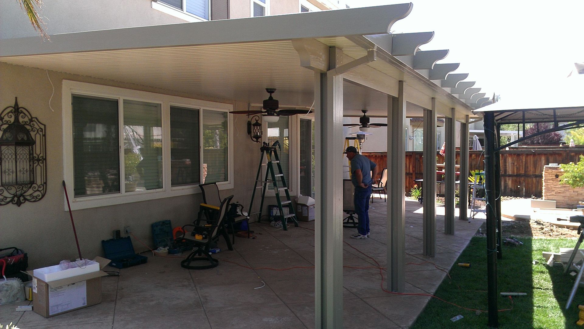 Covered backyard patio with white pergola, tools, and two people working near the house.