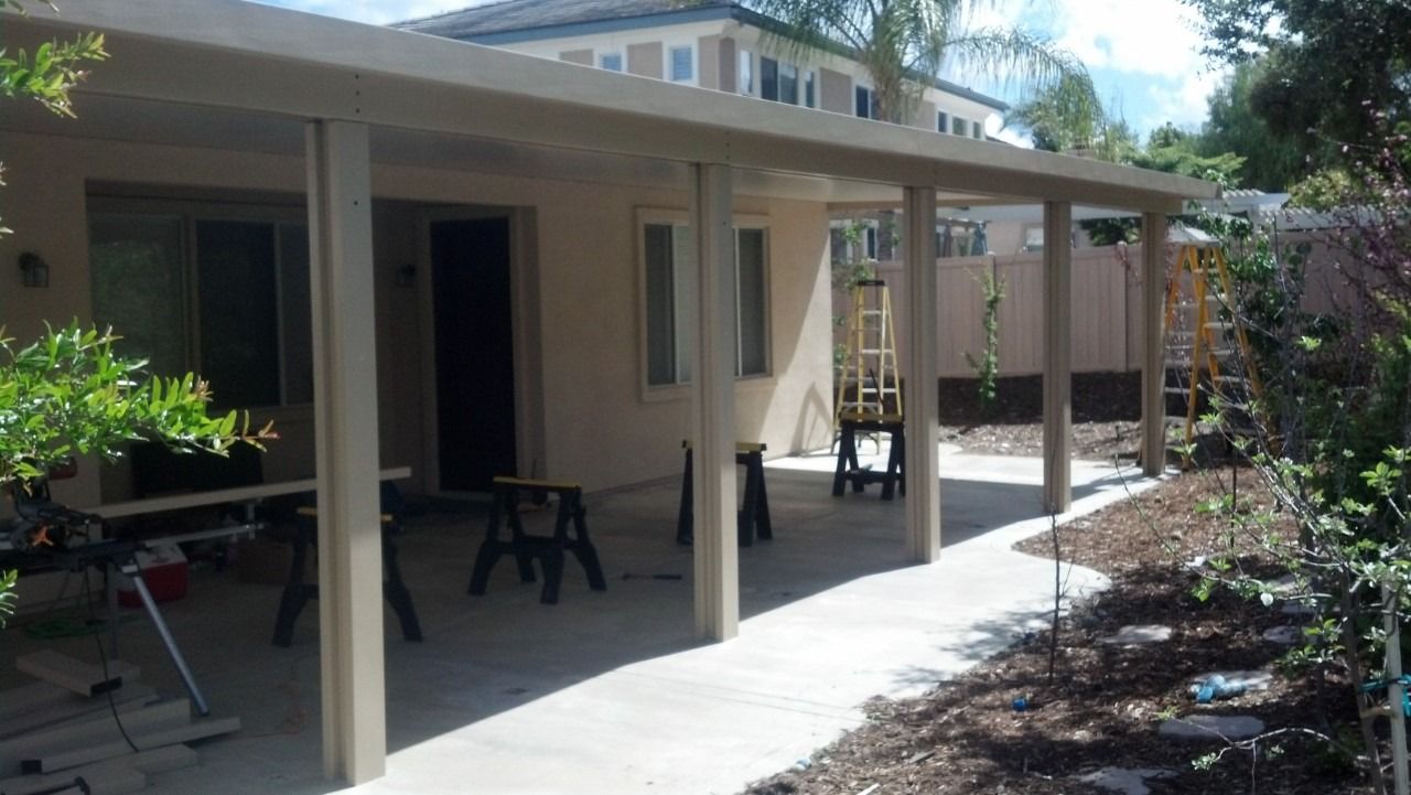Covered patio walkway beside a house, with white columns, shaded seating, and garden beds.