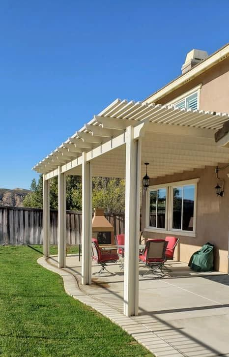 Back patio with white pergola, red chairs, beige house, and green lawn on a sunny day