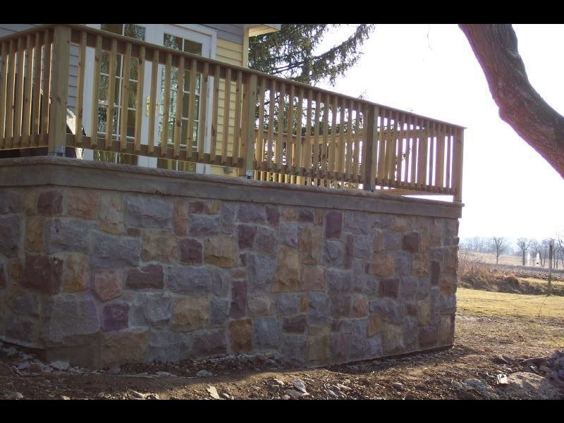 Stone foundation with wooden deck railing on a house, viewed outdoors in a sunny yard
