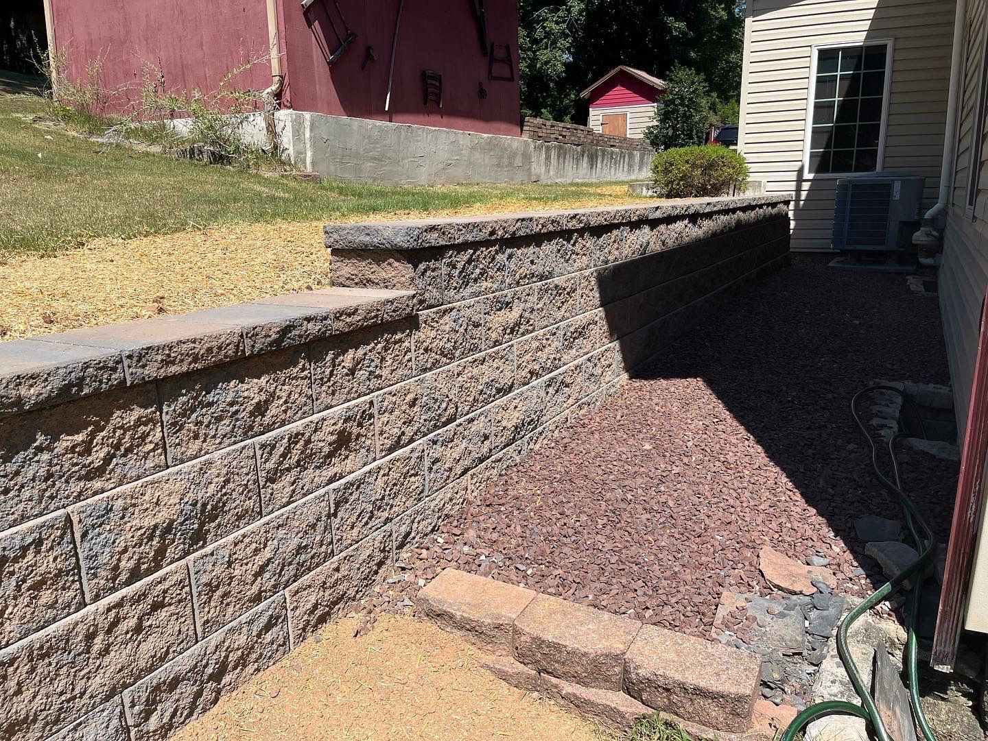 Stone retaining wall beside a house, with gravel walkway and narrow shaded side yard.