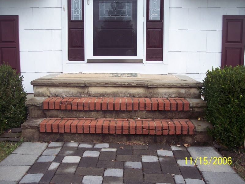 Front steps with red brick risers and stone landing leading to a white house entrance, flanked by shrubs.