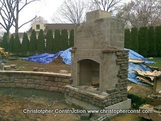 Stone outdoor fireplace under construction in a backyard, with blue tarps and stacked materials nearby