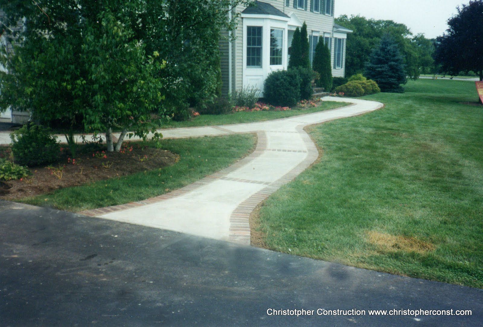 Curved concrete walkway through green lawn leading to a white house with trees and shrubs