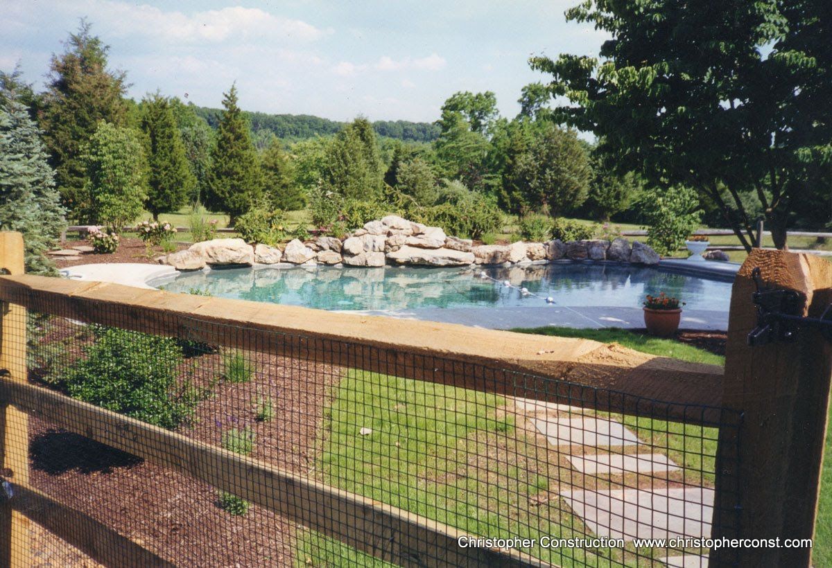 Backyard pool with rock waterfall, wooden deck, and lush green trees under a blue sky